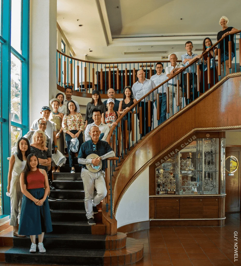 Group of people smiling and posing together on a curved staircase inside a building with large windows and wooden handrails. Some are standing on the steps, others on the upper landing. Display case and Marina Club sign visible.