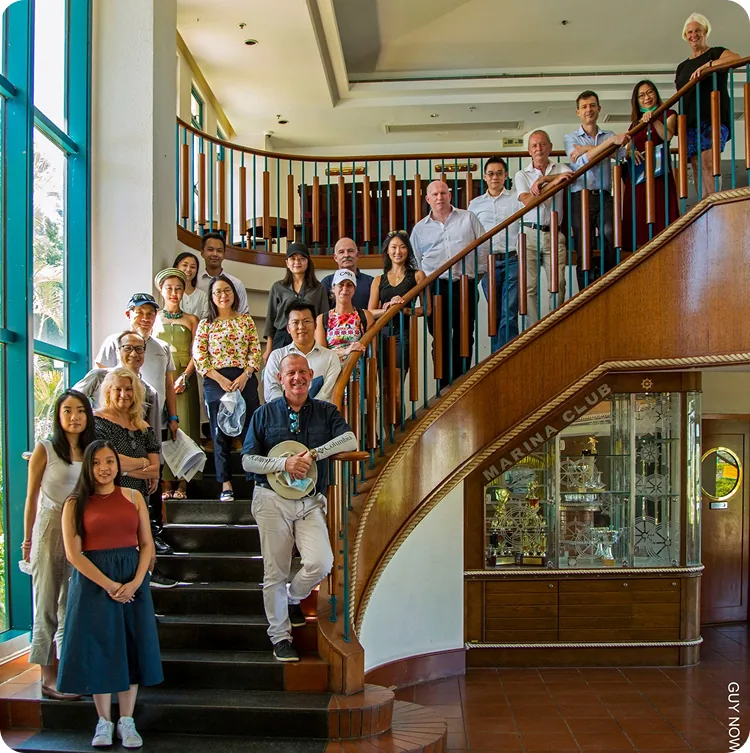 A diverse group of people stand and smile on a curved wooden staircase and at its base inside a bright, spacious room with large windows and a glass display case.