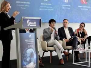 A woman speaks at a lectern while four panellists, three men and one woman, sit on stage with microphones and name cards behind them, participating in a conference discussion.