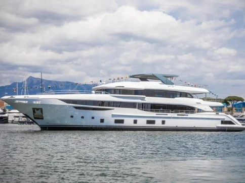 A large, modern white yacht with multiple decks is moored in a marina on a cloudy day, with mountains and other boats visible in the background.