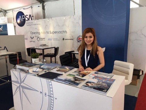 A woman with long hair is standing behind a stand at an event. The stand has brochures and magazines on display. Behind her is a banner reading APSA and Opening Asia to Superyachts, with several logos visible.
