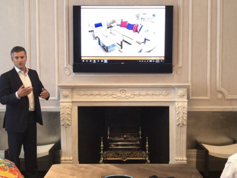 A man in a suit stands next to a fireplace, gesturing whilst presenting. Above the fireplace, a wall-mounted screen displays an image of a modern living room with sofas and colourful cushions.