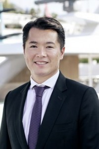A man in a dark suit, white shirt, and purple tie is smiling at the camera. The background is blurred, showing an outdoor setting with modern architecture.