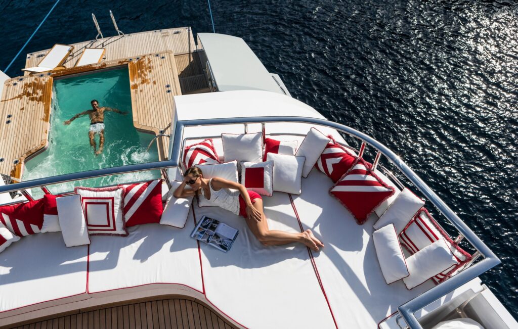 A woman lounges on a white and red-cushioned deck of a yacht, with a magazine beside her. Nearby, a man relaxes in a small pool at the yacht’s stern, surrounded by blue ocean water.