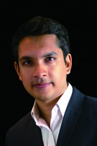 A man with short dark hair, wearing a dark pinstripe suit jacket and a white collared shirt, poses against a black background.