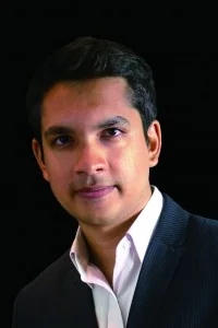 A man with short dark hair, wearing a dark pinstripe suit jacket and a white collared shirt, poses against a black background.