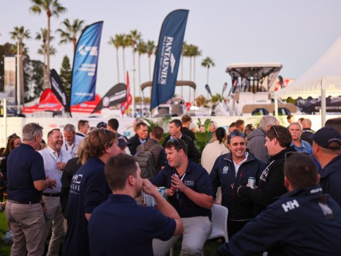 A group of people in navy shirts socialise outdoors at an event with flags, marquees, and boats in the background. Palm trees and a clear sky suggest a warm, coastal location.