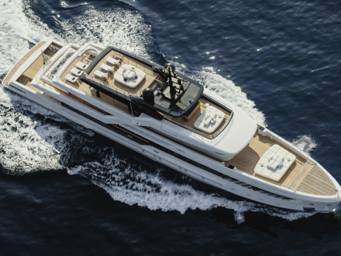 Aerial view of a large, modern white yacht cruising through deep blue water, leaving a trail of waves behind, with multiple deck areas furnished with seating and sun loungers.