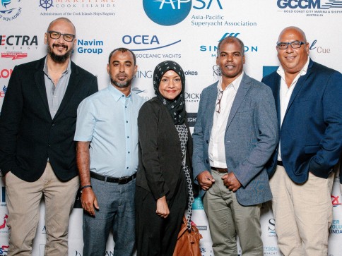Five people stand together smiling in front of a white backdrop featuring various company logos at an event. Four men wear business attire, and a woman in a black hijab stands in the middle.