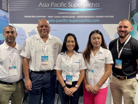Five people stand and smile in front of an Asia Pacific Superyachts stand display at an event. They are wearing name badges and branded shirts, posing together for a group photo.