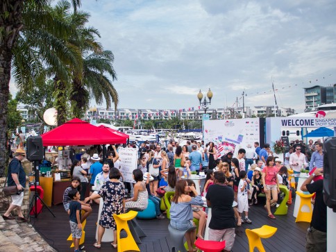 A lively outdoor event by the water, with people of all ages socialising and sitting on colourful stools. Palm trees, boats, banners, and a WELCOME sign are visible in the background under a cloudy sky.