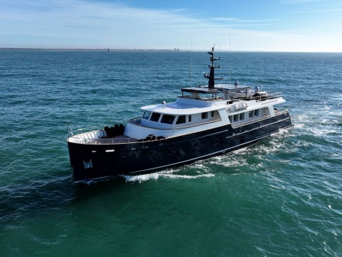 A modern motor yacht with a dark hull and white superstructure cruises through calm blue-green ocean water under a clear blue sky.