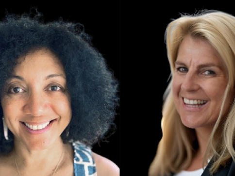 Two women smiling against a black background; the woman on the left has curly dark hair and wears a patterned top, whilst the woman on the right has long blonde hair and wears a dark top.