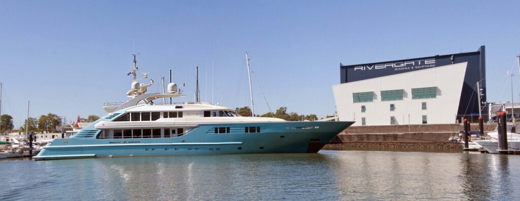 A large turquoise luxury yacht is docked in a harbour near a modern white building labelled Rivergate Marina & Shipyard under a clear blue sky.