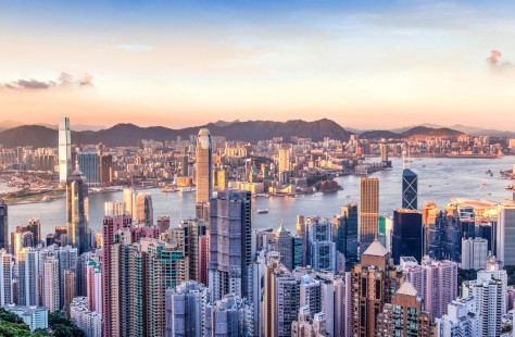 A panoramic view of Hong Kong’s skyline at sunset, featuring tall skyscrapers, Victoria Harbour, boats on the water, and distant mountains under a pastel sky.