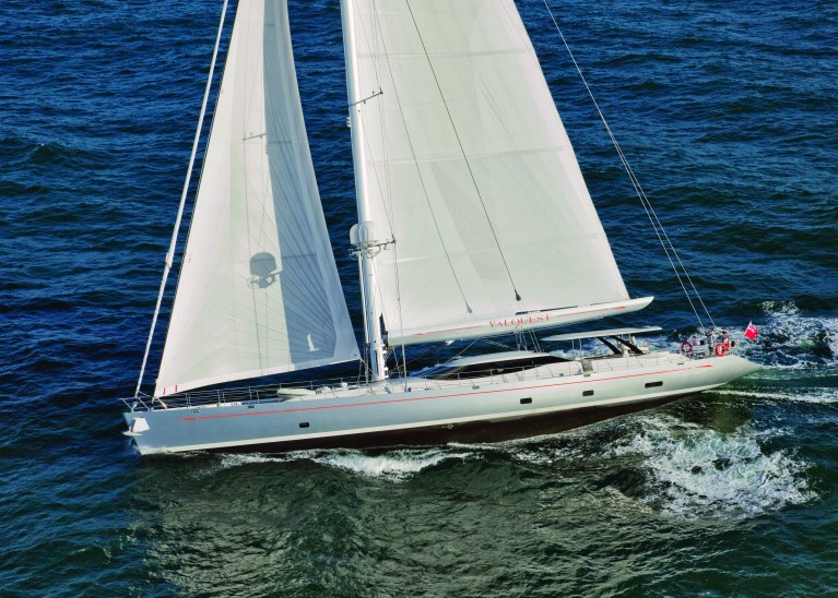 A large white sailing yacht with two masts glides across blue ocean waters, creating small waves as it moves. Several people are visible on the deck, and a red flag is flying at the stern.