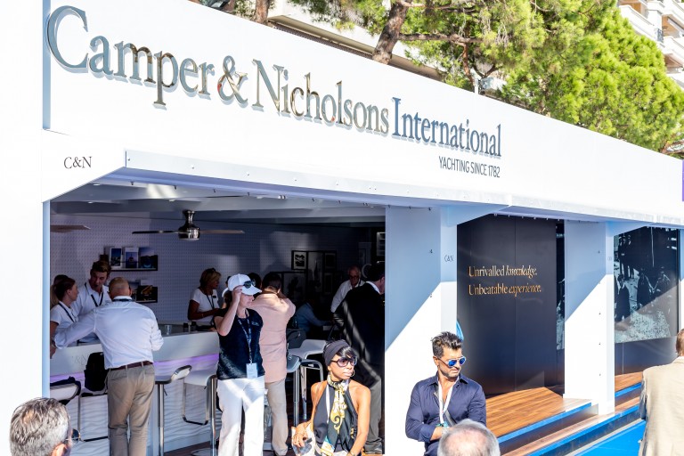 A group of people gather outside and inside a modern white Camper & Nicholsons International yachting stand, with large company signage and trees in the background. Some guests are talking, and others are seated at the bar.