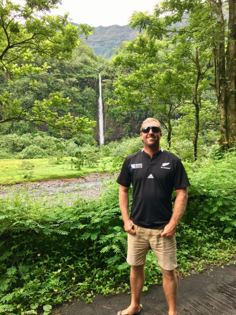 A man in sunglasses and a black shirt stands on a lush, green path with a tall waterfall and forested hills in the background.