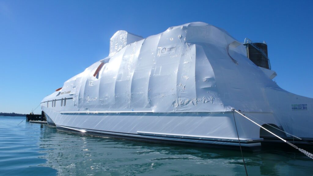 A large yacht moored in calm water is completely wrapped in white plastic shrink wrap, likely for protection or storage, under a clear blue sky.