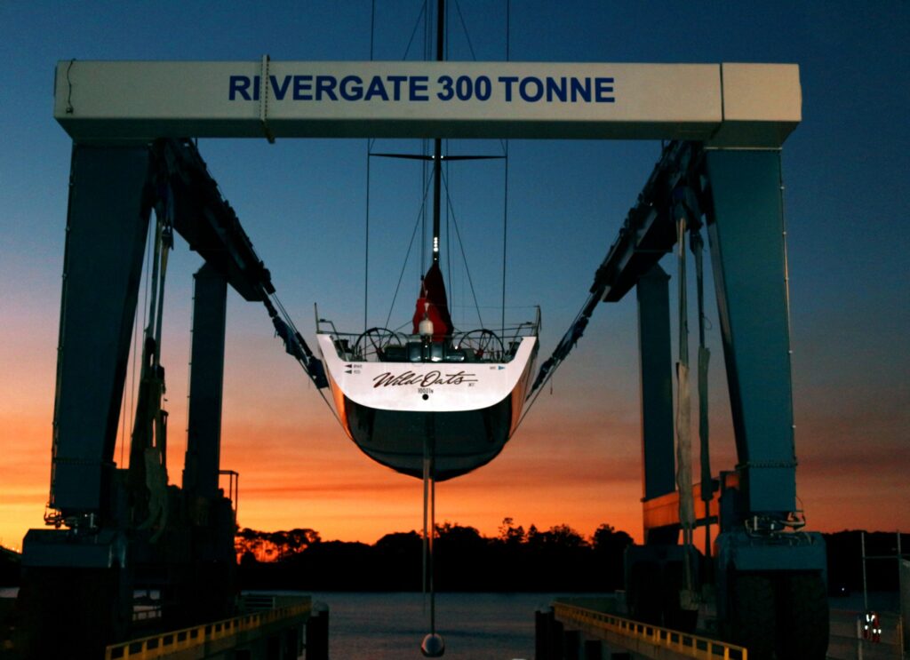 A sailboat is suspended on a boat lift labelled RIVERGATE 300 TONNE at sunset, with a vibrant orange sky and water in the background.