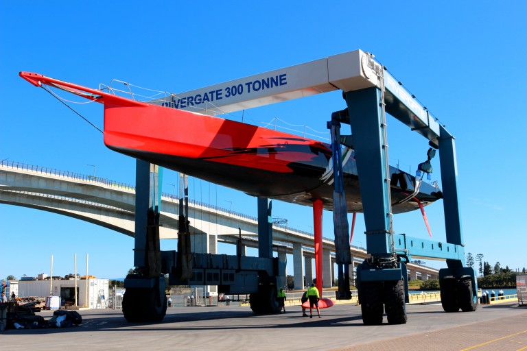 A large red sailboat suspended by a blue and white boat lift labelled WINDERCATE 300 TONNE at a marina, with a person in a green shirt working nearby under a clear blue sky.