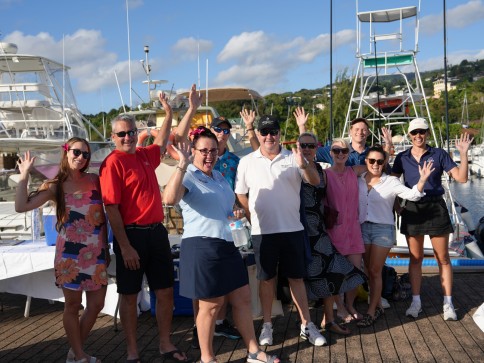 A group of people stand on a dock near boats, smiling and waving at the camera on a sunny day. Most are wearing sunglasses and casual summer clothes. Blue sky with some clouds is in the background.