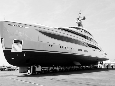 A large yacht is positioned on supports at a shipyard, appearing to be under construction or maintenance. The photo is in black and white, highlighting the vessel's sleek design and smooth contours.
