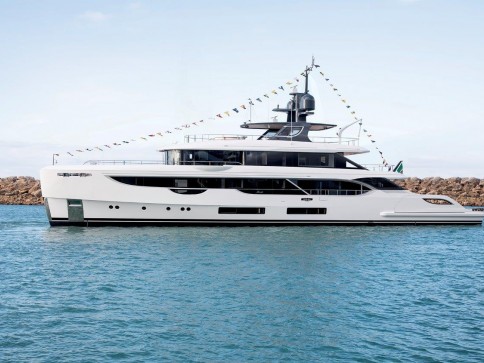 A sleek white luxury yacht decorated with colourful flags is anchored on calm blue water near a rocky breakwater under a clear sky.