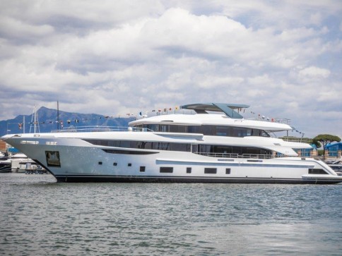 A large, modern white yacht with multiple decks is moored on calm water under a partly cloudy sky, with mountains visible in the background.