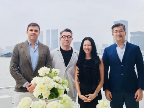 Four people stand together, dressed in smart casual and formal attire, smiling in front of a city skyline and waterfront. A table with white flowers is in the foreground.