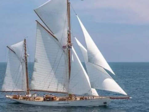 A large white yacht with multiple sails unfurled glides across calm blue sea water under a partly cloudy sky. Several people are visible on the deck.