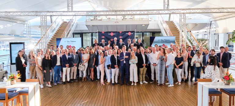 A large group of professionally dressed people pose together for a group photo on a wooden decking in an airy, well-lit venue with stairs, screens, and nautical decor in the background.
