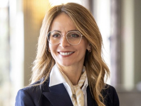 A woman with long, wavy blonde hair wearing glasses, a navy blazer, a white shirt, and a cream-coloured scarf smiles whilst standing indoors in a brightly lit setting.