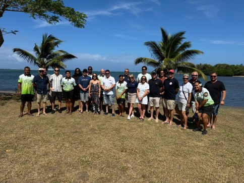 A group of about 22 people pose together outdoors near palm trees and water on a sunny day, standing on grass with a blue sky and sea in the background.