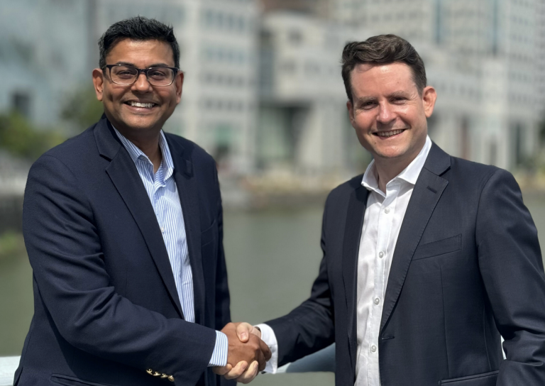 Two men in business suits are smiling and shaking hands outdoors, with a blurred cityscape and water in the background.