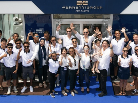 A large group of people in white shirts and sunglasses smile and wave outside a building with a Ferretti Group Asia Pacific sign above the entrance. They stand on a blue carpet in the sunshine.