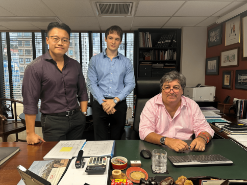 Three men in business attire are in an office. Two stand beside a desk, one in a dark shirt and the other in a light blue shirt; a third, in a pink shirt, sits at the desk with a keyboard. Office supplies are on the desk.