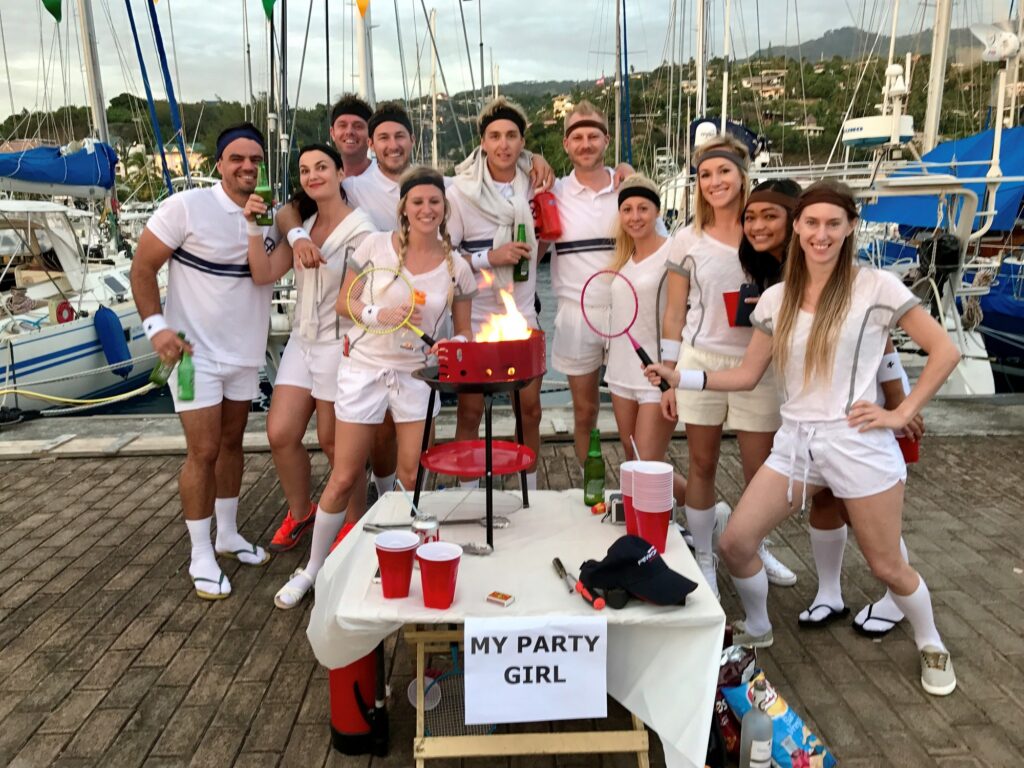 A group of people dressed in white tennis outfits pose and smile by a marina, standing around a barbecue and a table with drinks and a MY PARTY GIRL sign. Boats and hills are visible in the background.