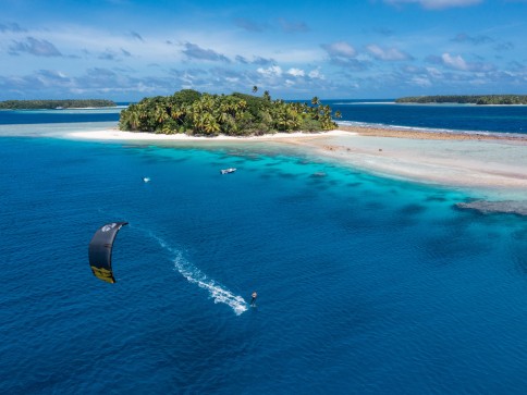 A person kitesurfing on clear blue water near a sandy, palm-covered tropical island, with a few boats anchored close to shore under a bright blue sky.