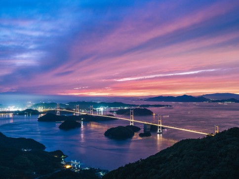 A long suspension bridge stretches across calm water between small islands at sunset, with vibrant purple and pink hues in the sky and city lights glowing in the distance.