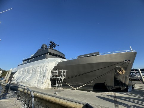 A large, modern yacht with a dark hull is moored at a marina. Part of the yacht is covered in white protective sheeting, and scaffolding stands nearby under a clear blue sky.