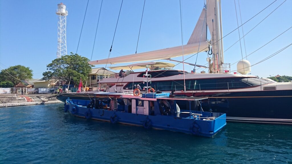 A large yacht with white sails berthed at a marina, with a smaller blue boat moored alongside. A building, trees, and a tall lattice tower are visible in the background under a clear blue sky.