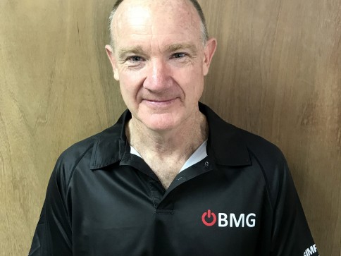 A middle-aged man with short hair is standing in front of a wooden wall, wearing a black collared shirt with the BMG logo on it. He is smiling slightly and facing the camera.