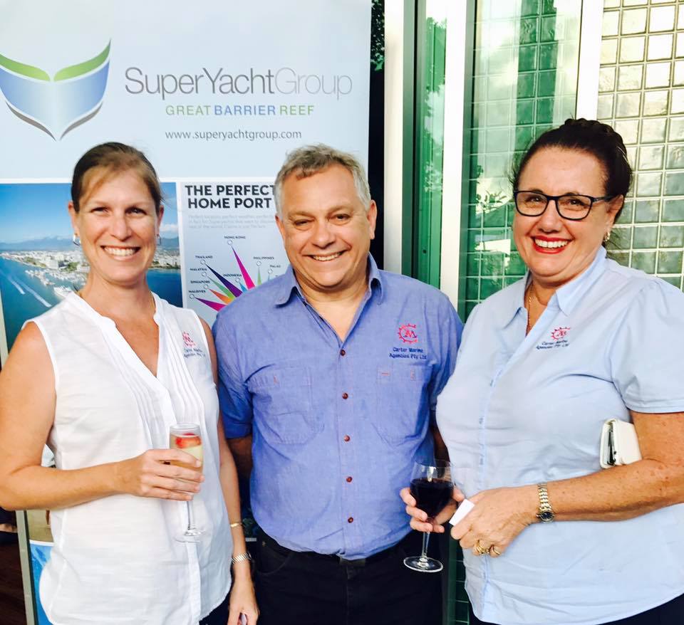 Three people stand smiling in front of a SuperYacht Group Great Barrier Reef banner. The two women hold glasses of wine, whilst the man stands between them. All are dressed in casual business attire.