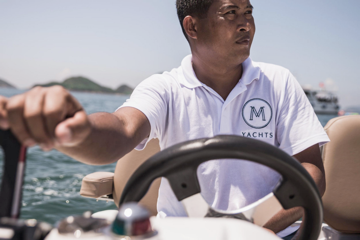 A man in a white YACHTS polo shirt is steering a boat on the water, looking to the side. The sunny sky and other boats are visible in the background.