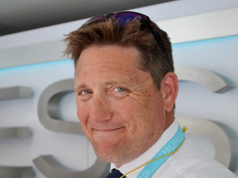 A man with short brown hair and freckles smiles at the camera. He is wearing a white shirt, blue tie, and a lanyard with BURGESS printed on it. Sunglasses rest on his head and a metallic sign is in the background.