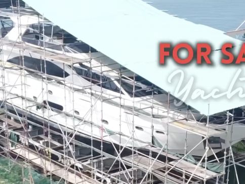 A large yacht under construction is covered by scaffolding and a canopy. Bold red text reads FOR SALE with Yacht in script below it. The sea is visible in the background.