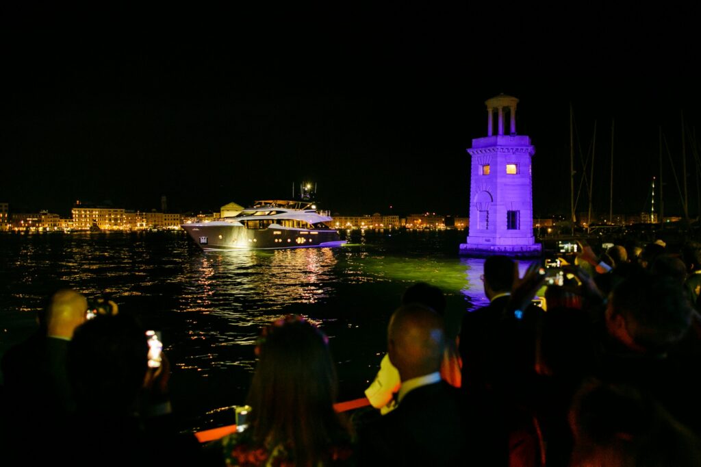 A crowd watches and takes photos as a yacht illuminated by lights sails near a small, tower-like structure glowing purple on the water at night, with city lights in the background.
