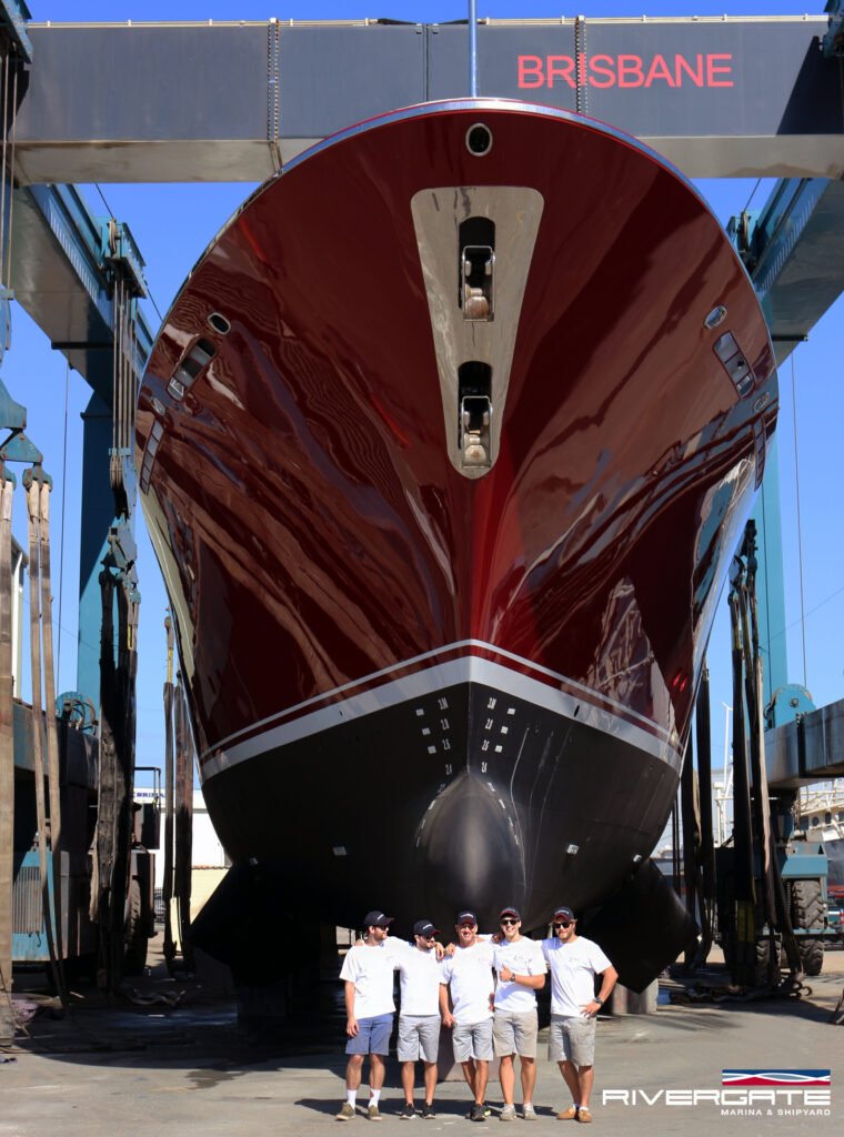 Five people in matching uniforms stand in front of a large, red and black boat in a shipyard. The boat is elevated on supports under a gantry crane labelled “BRISBANE.” The Rivergate logo is visible in the corner.