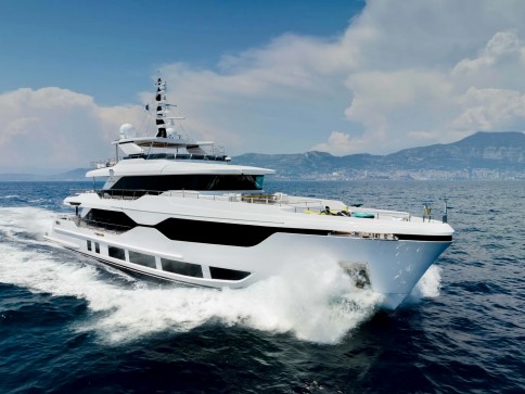 A large white luxury yacht cruises through calm blue ocean waters, creating a wake. The sky is mostly clear with some clouds, and distant mountains are visible on the horizon.